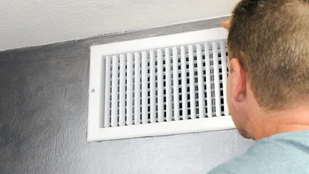 Man inspecting a white air vent on a gray wall, focusing on ventilation system maintenance or repair.