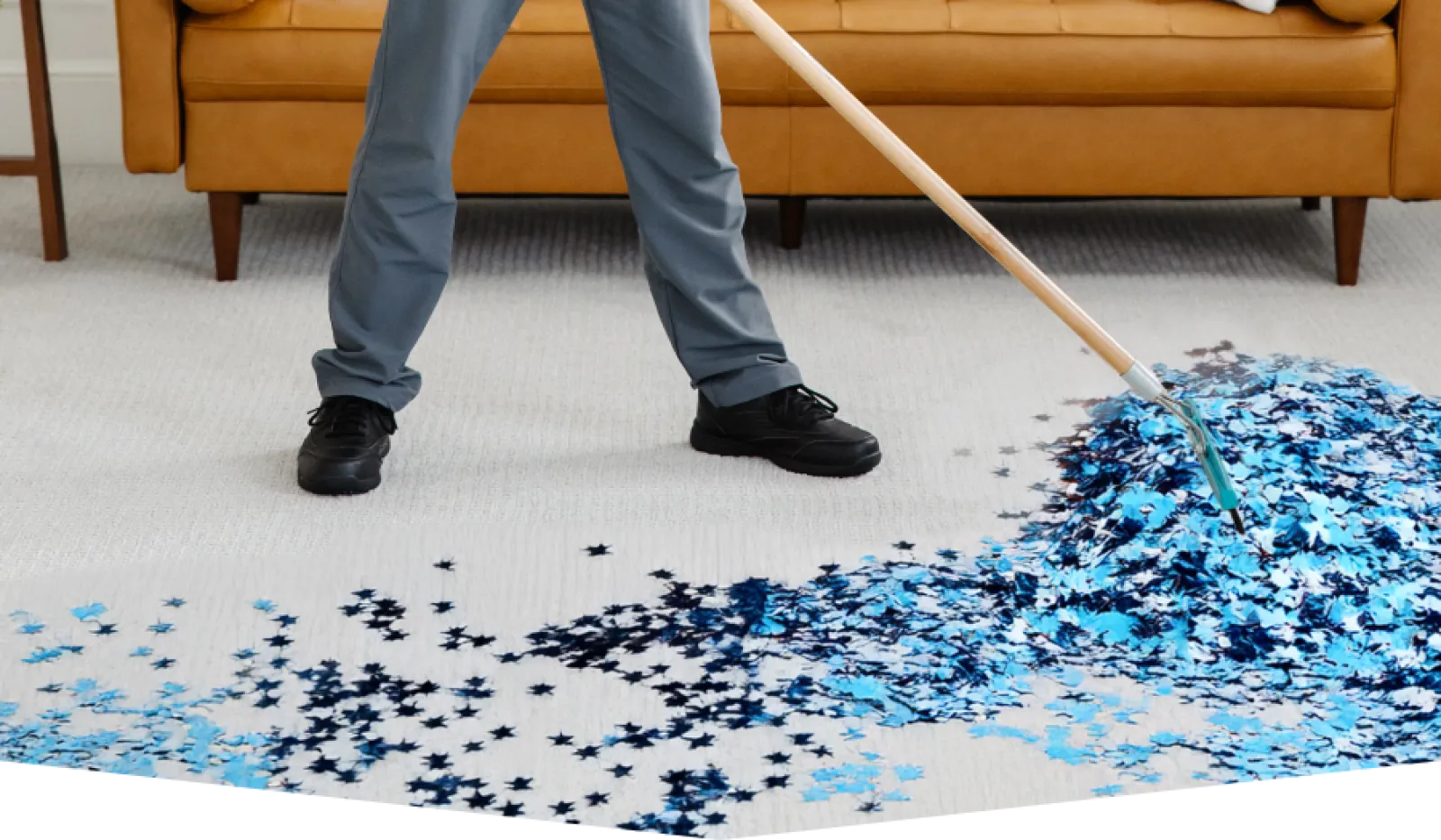 Person using a broom to sweep blue and black star-shaped confetti on a light carpet in front of a brown sofa.