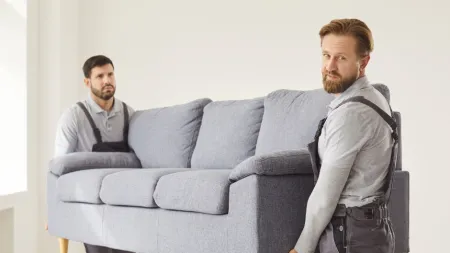 Two movers carrying a gray sofa in a bright, minimalist room with wooden flooring and white walls.